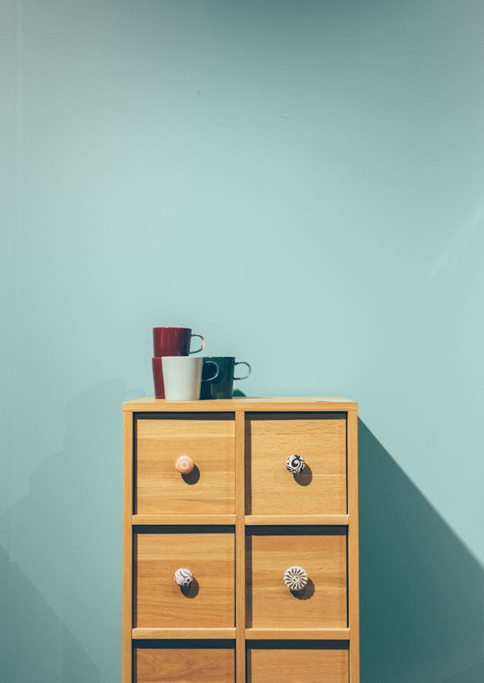 A stylish wooden cabinet topped with assorted mugs against a pastel wall background.